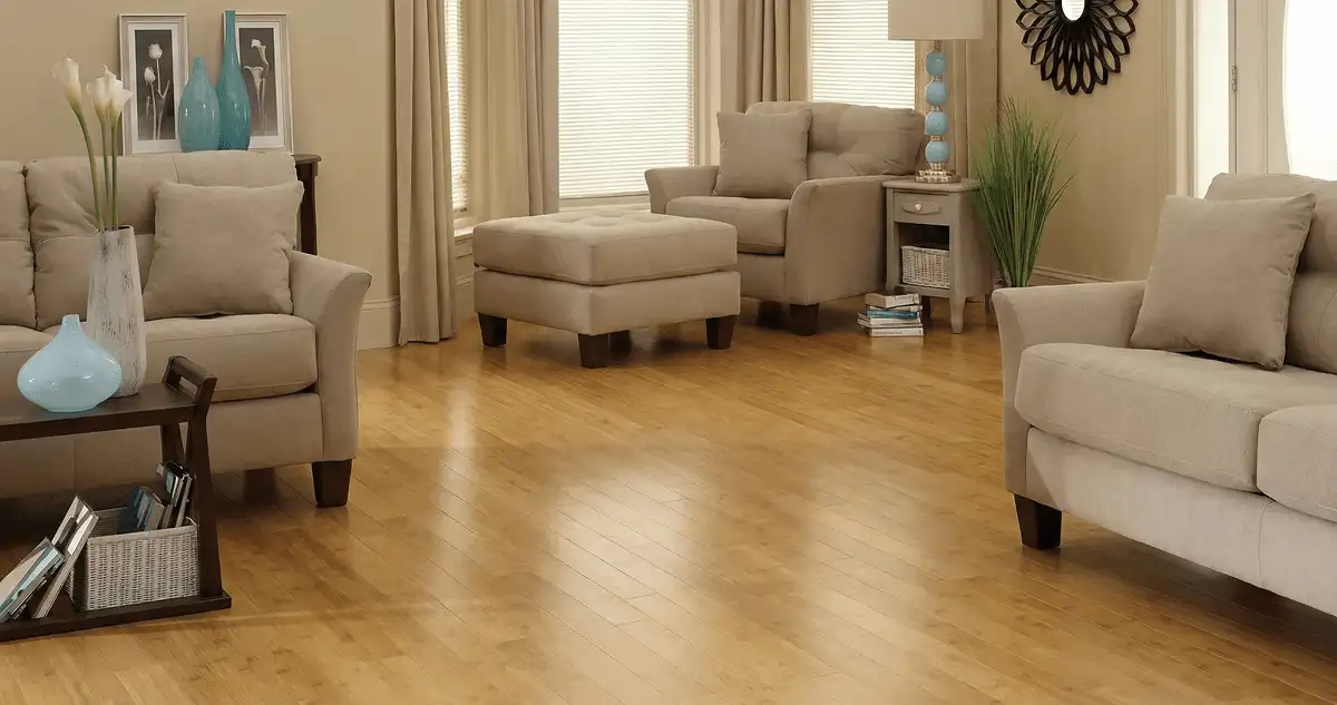A living room with professionally installed bamboo flooring, beige sofas, a matching ottoman, a side table with a blue lamp, vases, framed pictures, and large windows with blinds and curtains in Duluth, MN